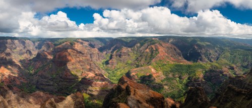 A panoramic view of a lush canyon with colorful rock formations and a blue sky dotted with clouds.