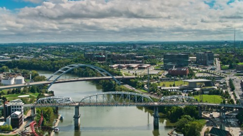 Aerial view of a cityscape featuring bridges over a river, green parks, and modern buildings under a cloudy sky.