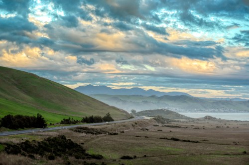 A scenic landscape featuring rolling green hills, a winding road, and mountains under a cloudy sky.