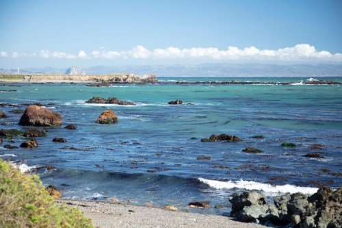 A serene coastal scene with rocky shores, clear blue water, and distant mountains under a bright sky.