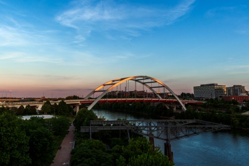 A modern bridge arches over a river, with lush greenery and buildings in the background under a blue sky.