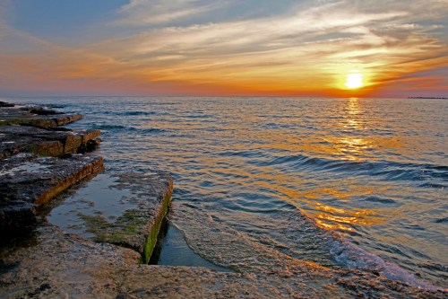 Sunset over calm waters, with rocky shoreline reflecting warm hues of orange and gold in the sky.