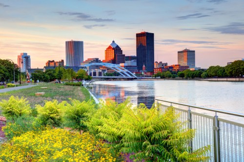 City skyline at sunset reflecting on a river, with lush greenery and flowers in the foreground.