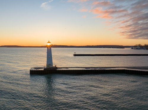 A lighthouse stands on a pier at sunset, casting light over calm waters and a colorful sky.