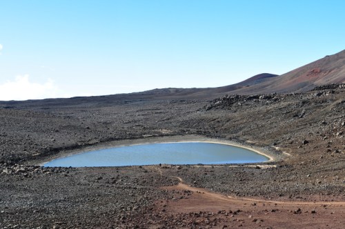 A serene, oval-shaped pond surrounded by rocky terrain and distant mountains under a clear blue sky.