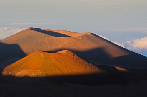 Sunlit volcanic landscape with two distinct mountains casting shadows against a backdrop of clouds.