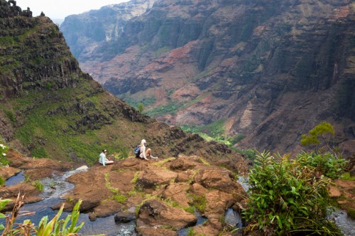 Two people sit on a rocky ledge overlooking a lush, mountainous valley with steep cliffs and vibrant greenery.
