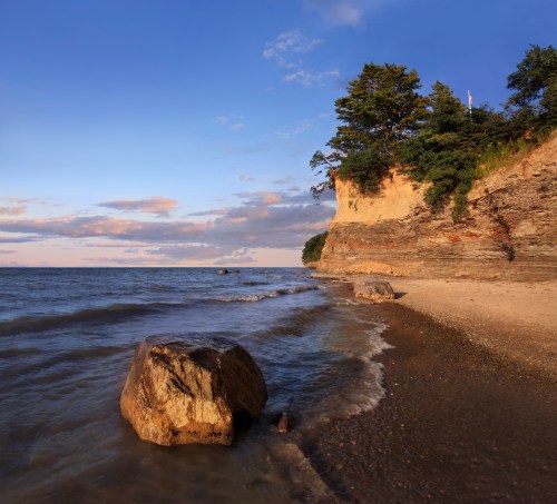 A serene beach scene with rocky shoreline, gentle waves, and a cliff with trees under a blue sky with scattered clouds.