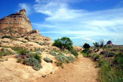 A scenic landscape featuring rocky formations, a dirt path, and sparse greenery under a blue sky with clouds.