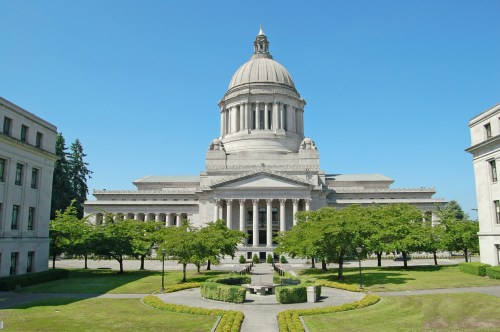 A grand government building with a large dome, surrounded by green trees and a manicured lawn under a clear blue sky.