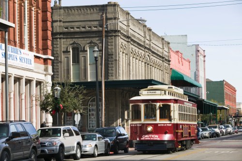 A vintage streetcar travels along a historic street lined with buildings and parked cars.