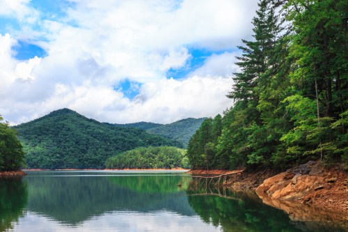 Serene lake surrounded by lush green mountains and trees under a partly cloudy sky. Reflections on the water's surface.