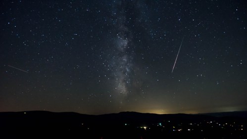 A starry night sky with the Milky Way and shooting stars over a dark landscape with distant lights.