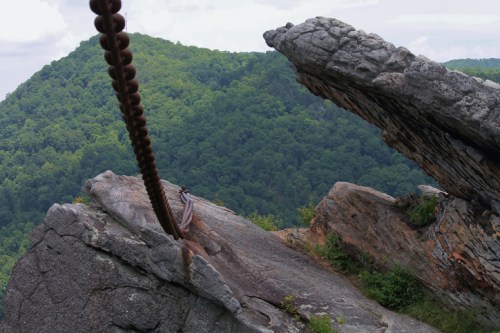 A rocky cliff with a chain extending over the edge, overlooking a lush green mountain landscape.