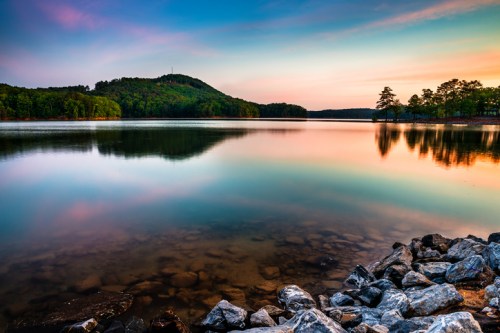 A serene lake at sunset, reflecting colorful skies and surrounded by lush green hills and rocky shoreline.