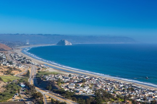 A scenic coastal view featuring a sandy beach, ocean waves, and a distant rock formation under a clear blue sky.