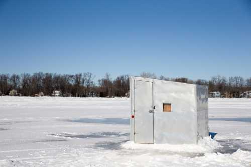 A metal ice fishing shelter on a snowy lake under a clear blue sky, surrounded by a frozen landscape.