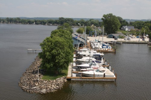 Aerial view of a marina with boats docked, surrounded by trees and calm water on a sunny day.