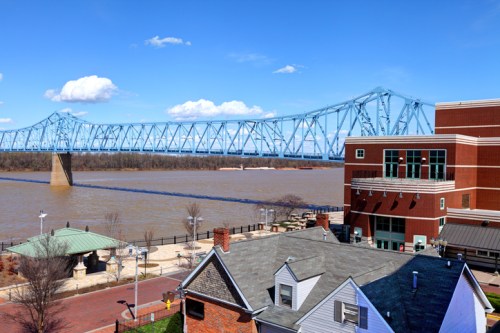 A blue bridge spans a river, with buildings and a park visible in the foreground under a clear blue sky.