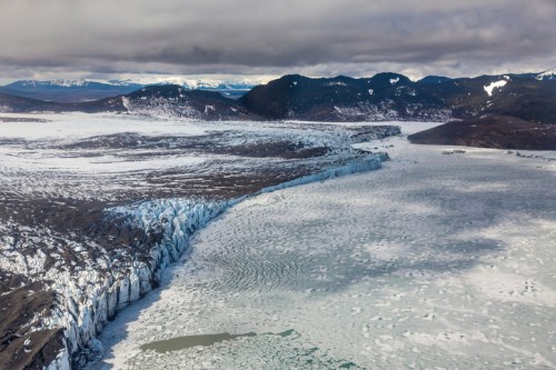 Aerial view of a glacier meeting a frozen lake, surrounded by mountains under a cloudy sky.