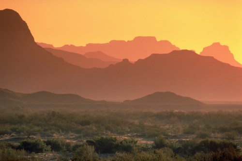 Silhouetted mountains against a vibrant orange sunset, with a foreground of sparse vegetation.