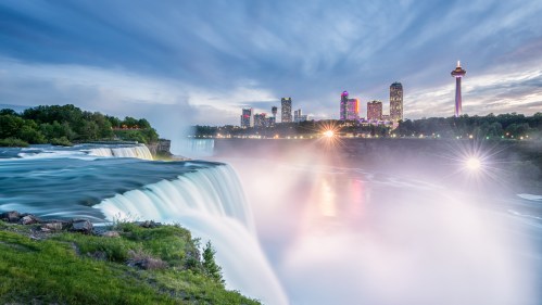 A panoramic view of Niagara Falls with a city skyline and colorful lights in the background under a cloudy sky.