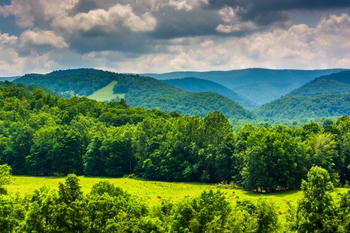Lush green hills and valleys under a cloudy sky, with trees and a grassy field in the foreground.