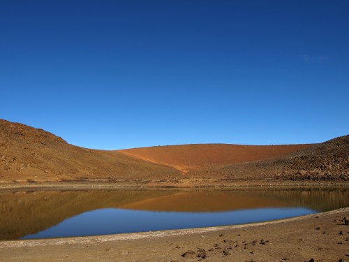 A serene landscape featuring a calm lake reflecting a clear blue sky, surrounded by rocky hills and dry terrain.