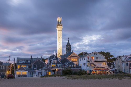 A tall lighthouse illuminated at dusk, surrounded by charming beach houses and a cloudy sky.