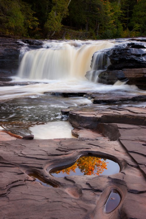 A serene waterfall cascades over rocks, with a small pool reflecting autumn foliage nearby.