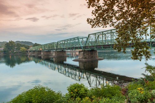 A green metal bridge spans a calm river, reflecting in the water, surrounded by trees and a cloudy sky.