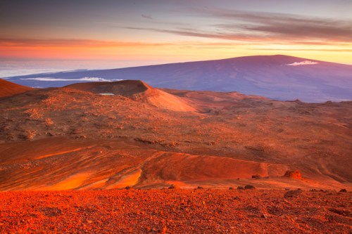 A stunning landscape of a volcanic terrain at sunset, with vibrant orange hues and a distant mountain silhouette.
