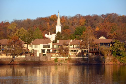 A serene lakeside view featuring autumn trees, a church steeple, and waterfront homes reflecting in the water.