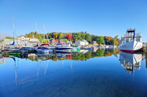 A serene harbor scene with colorful boats reflecting in calm water, surrounded by trees in autumn foliage.