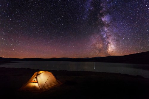 A glowing tent by a lake under a starry sky, showcasing the Milky Way and distant mountains.