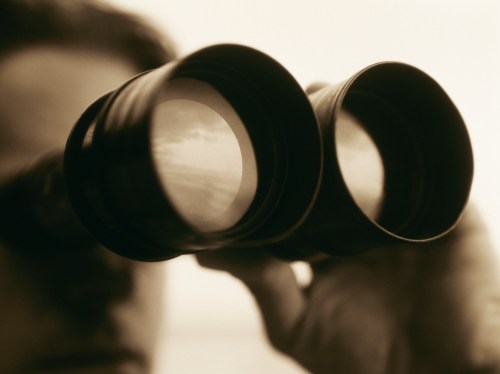 A person holding binoculars, looking through them with a blurred background, suggesting observation or exploration.