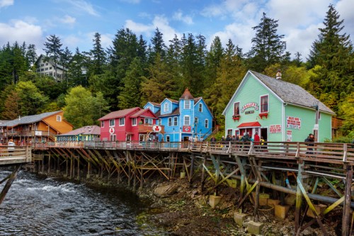 Colorful buildings on stilts by a river, surrounded by trees and a cloudy sky. A vibrant waterfront scene.