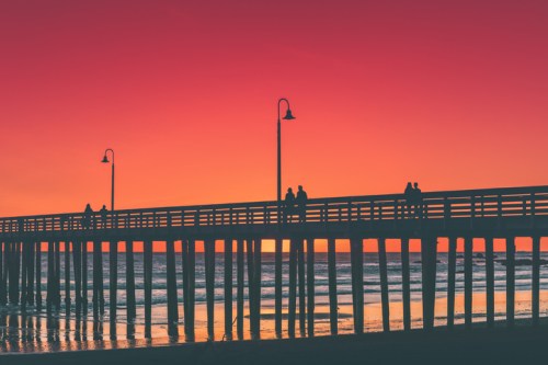 Silhouetted pier at sunset, with people walking and vibrant orange and pink sky reflecting on the water.