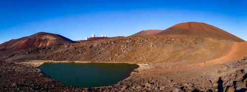 A panoramic view of a volcanic landscape with a small lake, rocky terrain, and observatories in the background.