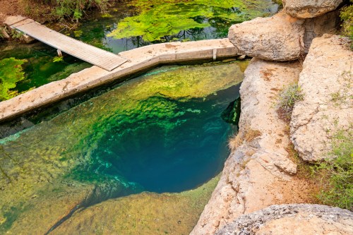 A clear, greenish-blue natural pool surrounded by rocks and vegetation, with a wooden bridge crossing over it.