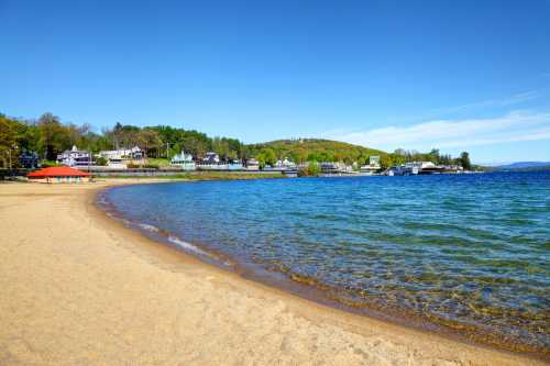 A sandy beach curves along a clear blue lake, with green hills and colorful houses in the background under a bright sky.