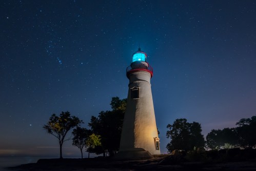 A lighthouse illuminated at night, surrounded by trees under a starry sky.