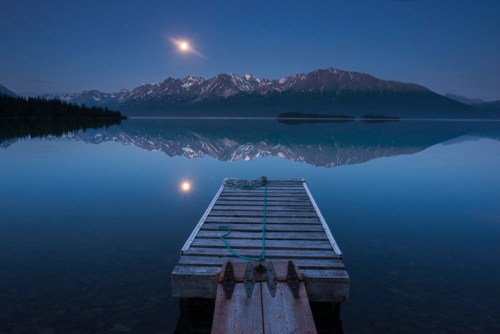 A serene lake at dusk with a wooden dock, mountains in the background, and a full moon reflecting on the water.