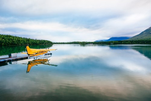 A yellow seaplane docked on a calm lake, surrounded by lush greenery and mountains under a cloudy sky.