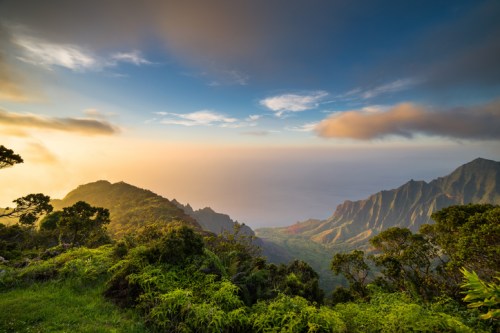 A scenic view of lush green mountains meeting the ocean under a colorful sky at sunset.