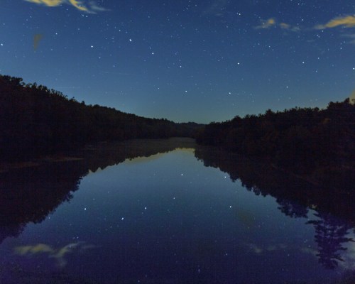 A serene night scene of a calm river reflecting a starry sky and surrounding trees.
