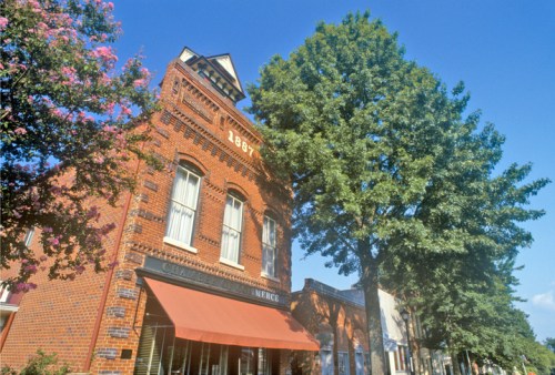 Historic brick building with "1887" on the facade, shaded by a large tree under a clear blue sky.