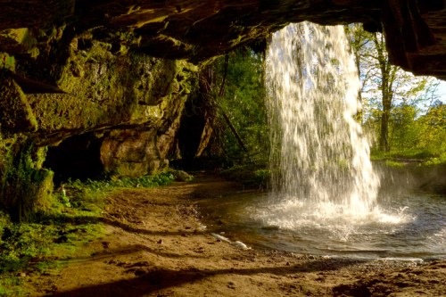 A serene waterfall cascades from a rocky overhang into a tranquil pool surrounded by lush greenery.