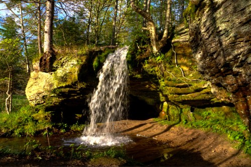 A serene waterfall cascades over rocky terrain, surrounded by lush greenery and trees in a peaceful forest setting.
