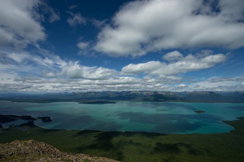 A panoramic view of a turquoise lake surrounded by lush greenery and mountains under a partly cloudy sky.